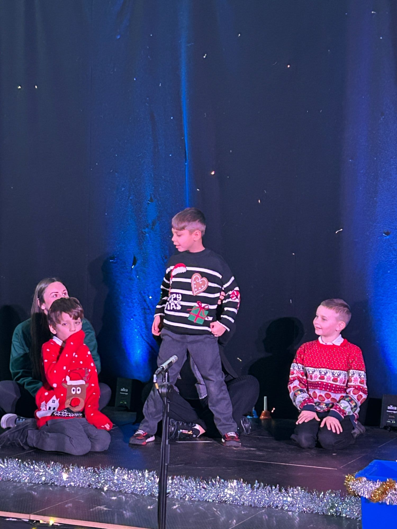 Two boys sitting on stage and one standing, all in Christmas jumpers