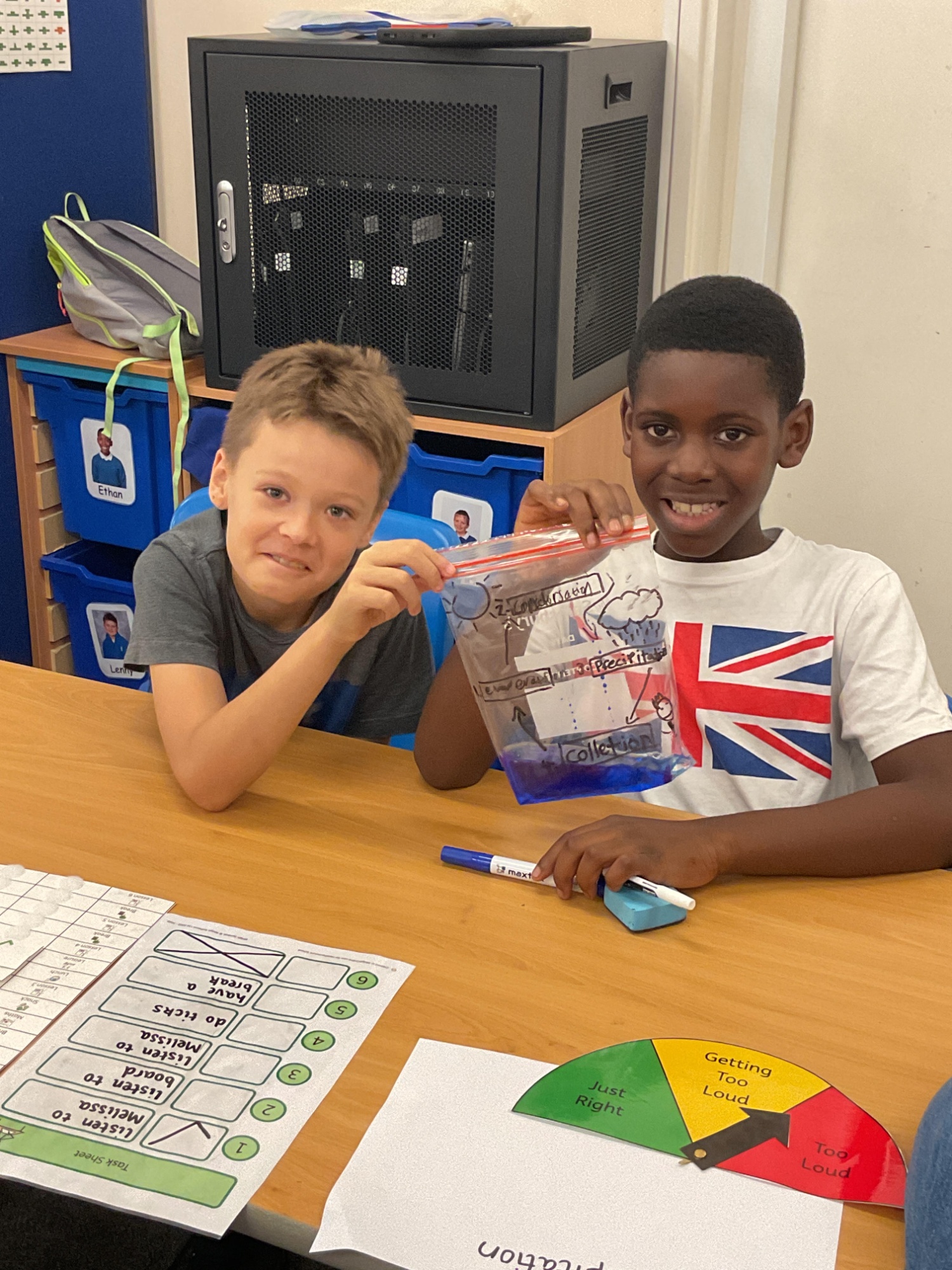 Two boys sat at a desk smiling holding up a bag of water