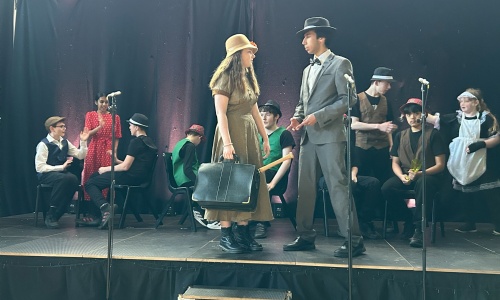 Blousy and Bugsy stood on stage at a bar scene with other cast members sat behind them