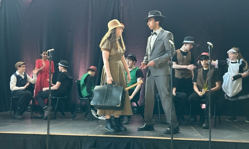 Blousy and Bugsy stood on stage at a bar scene with other cast members sat behind them