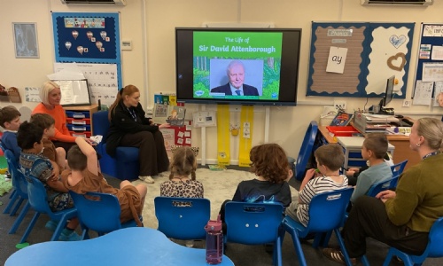EYFS students sitting in a semi circle watching a presentation on the life of David Attenborough.