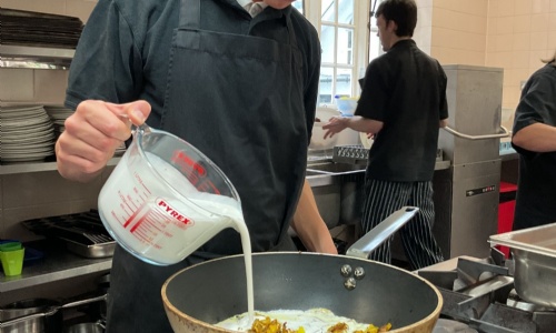 Student pouring coconut milk into his wok whilst cooking his curry dish