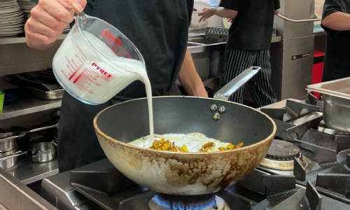 Student pouring coconut milk into his wok whilst cooking his curry dish
