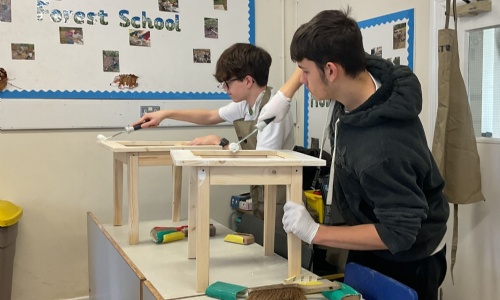 Two construction students working side by side painting their side tables with a roller brush